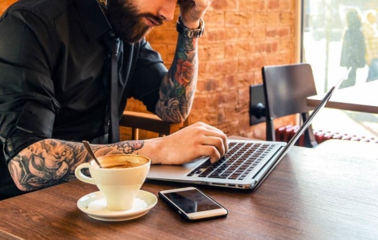 Man with beard and tattoos working on laptop in a cafe