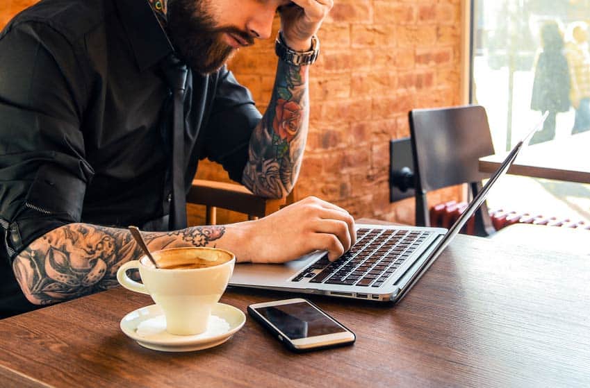 Man with beard and tattoos working on laptop in a cafe