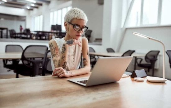 Tattooed Booking Agent in eyeglasses looking at laptop screen while working in modern working space