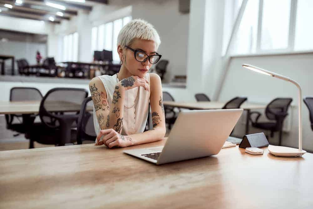 Tattooed Booking Agent in eyeglasses looking at laptop screen while working in modern working space