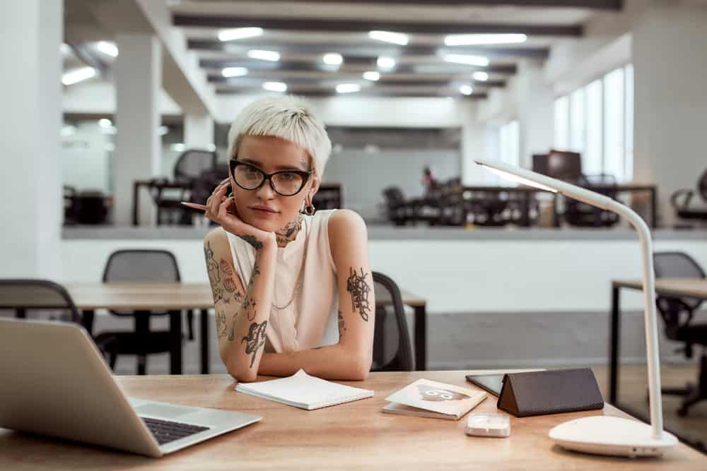 Pretty and young tattooed musician in eyewear looking at camera while working alone in the modern office