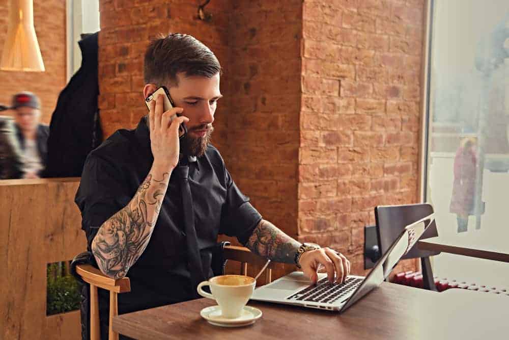 Stylish bearded hipster musician talking by smartphone and using a laptop in a coffee shop