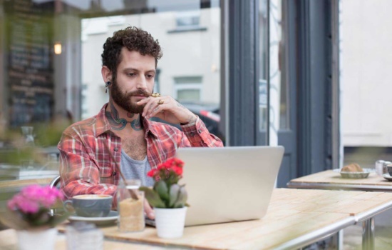 Tattooed male musician looking up music marketing strategies on his laptop at an outdoor cafe