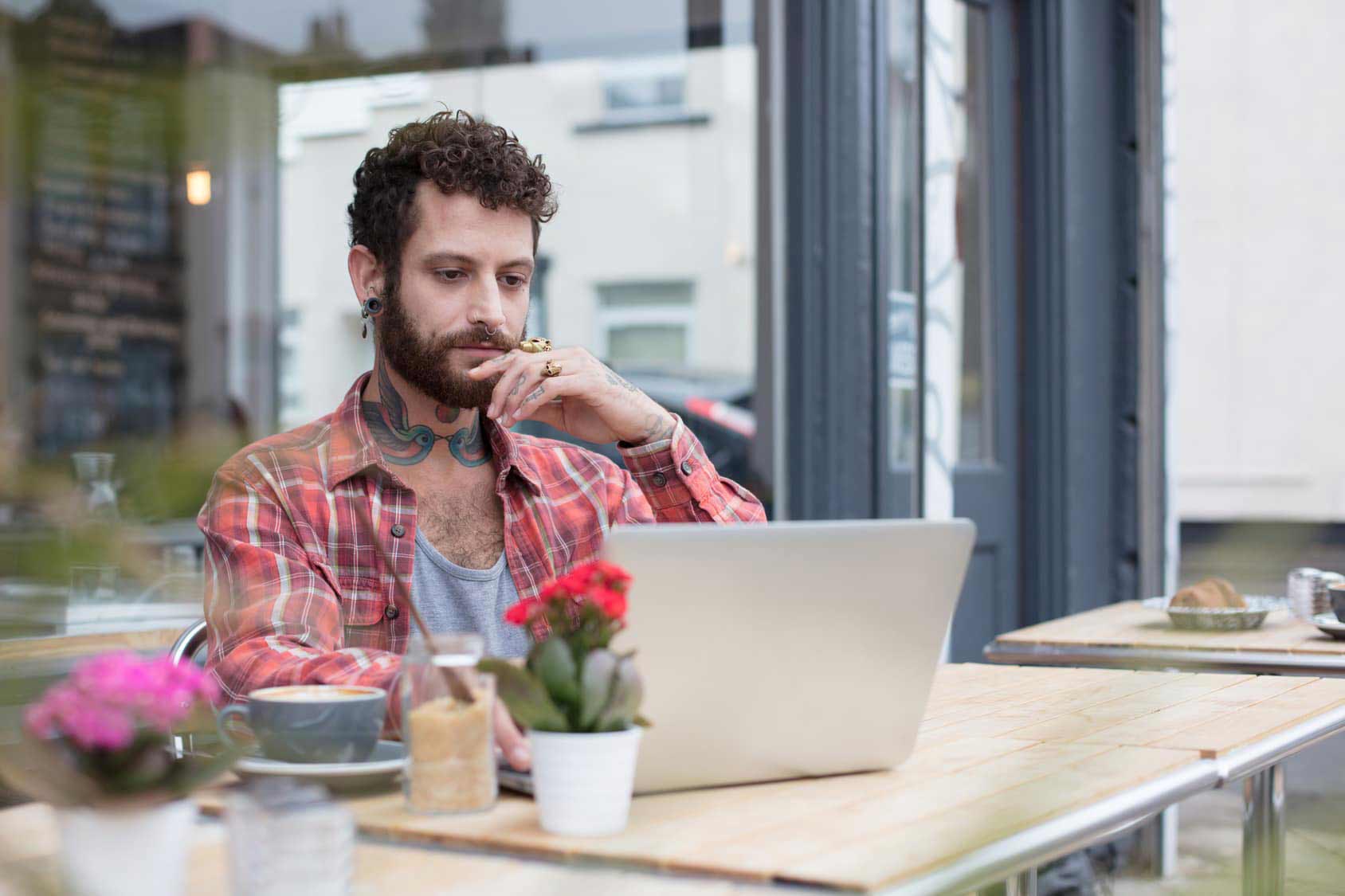 Tattooed male musician looking up music marketing strategies on his laptop at an outdoor cafe