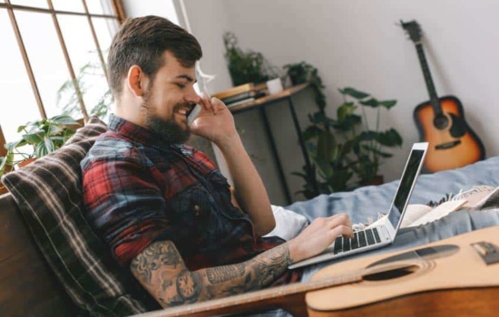 Tattooed male guitar player talking on the phone as he looks at his laptop