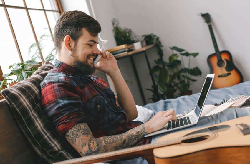 Tattooed male guitar player talking on the phone as he looks at his laptop