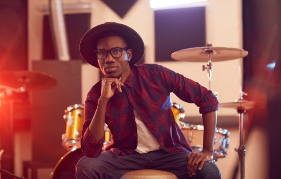 Musician in hat and plaid shirt sitting with drum kit in rehearsal studio