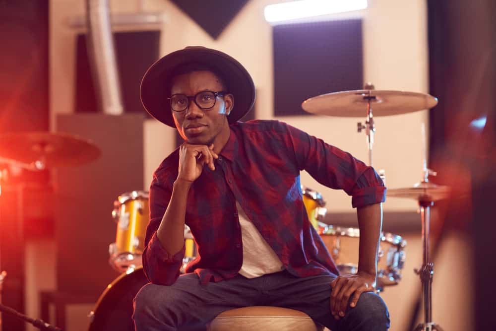 Musician in hat and plaid shirt sitting with drum kit in rehearsal studio