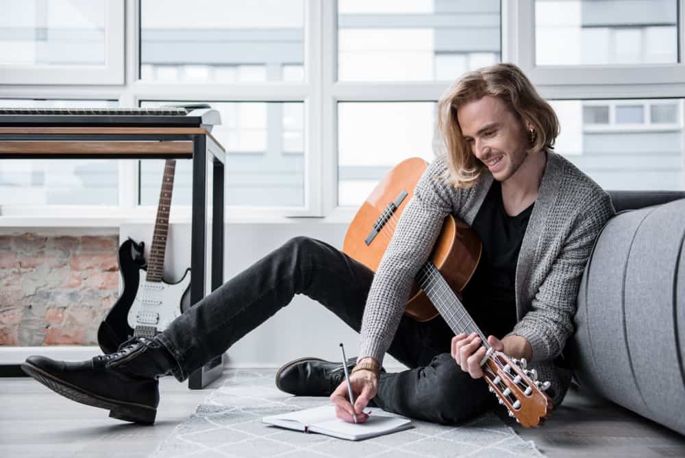 Young male musician sitting on floor with guitar and lyrics notebook