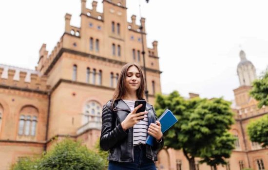 Young female student in leather jacket checking smart phone on college campus
