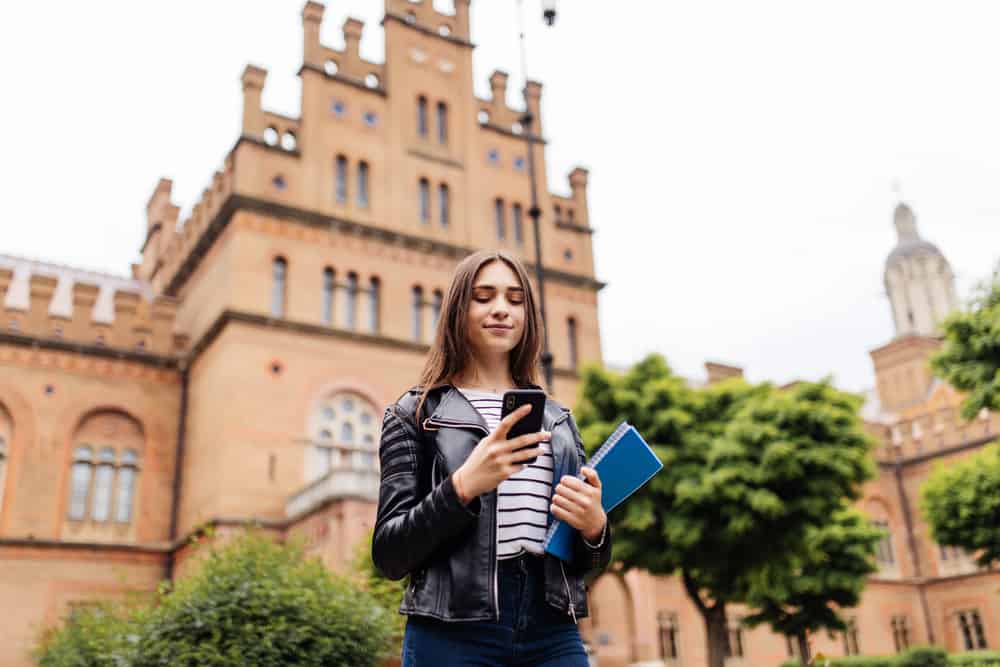 Young female student in leather jacket checking smart phone on college campus