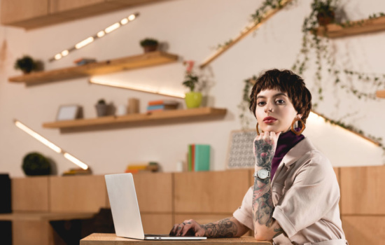 Tattooed staff publicist working on her laptop in large, modern office space